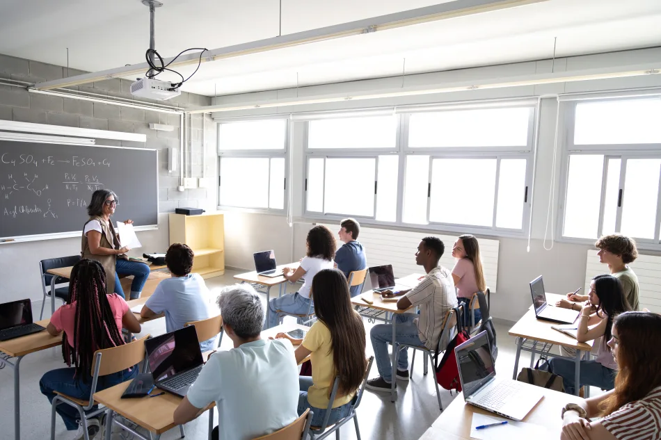 Un classe di studenti, ripresa di spalle e seduta ai propri banchi, ascolta la lezione di una professoressa