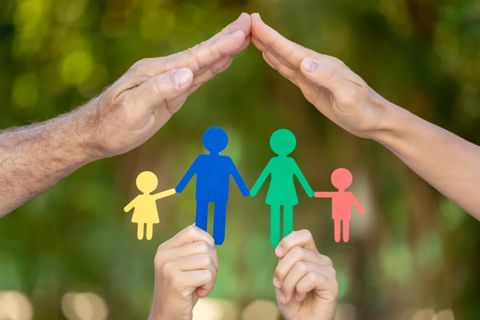 Due mani di adulto si uniscono a formare un tetto protettivo per una serie di figurine di carta colorate rappresentanti una famiglia, sostenute dalle mani di un bambino