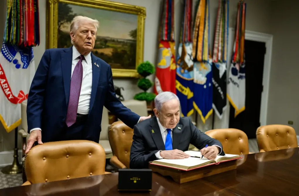 Donald Trump, in piedi, poggia una mano sulla spalla di Benjamin Netanyahou, impegnato nella firma della tregua con Hamas
