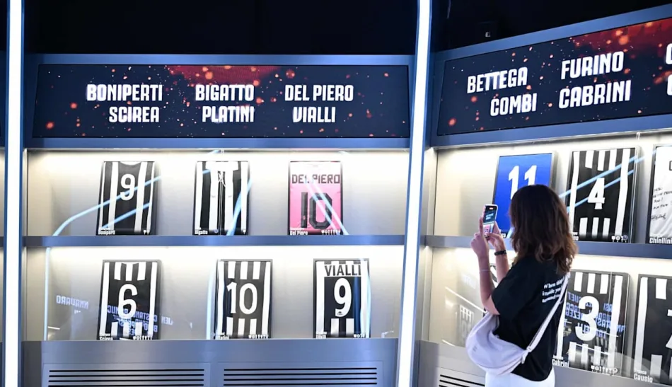 Una galleria di maglie di celebri campioni della Juventus, da Del Piero a Scirea, da Combi a Causio, da Boniperti a Platinì. All'interno, una visitatrice scatta una fotografia.