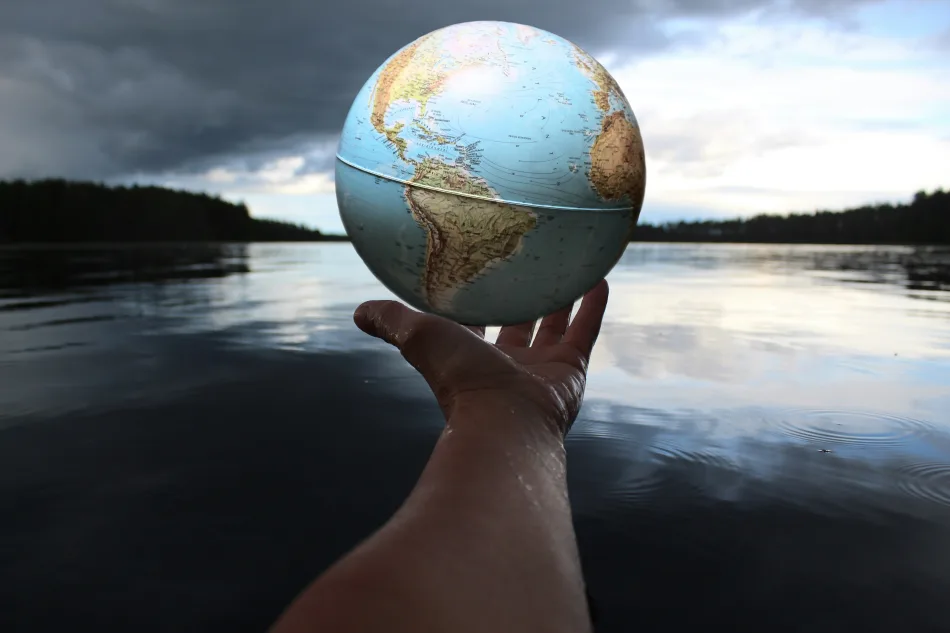 Un globo terrestre tenuto sulle dita di una mano con sullo sfondo un bacino di acqua.