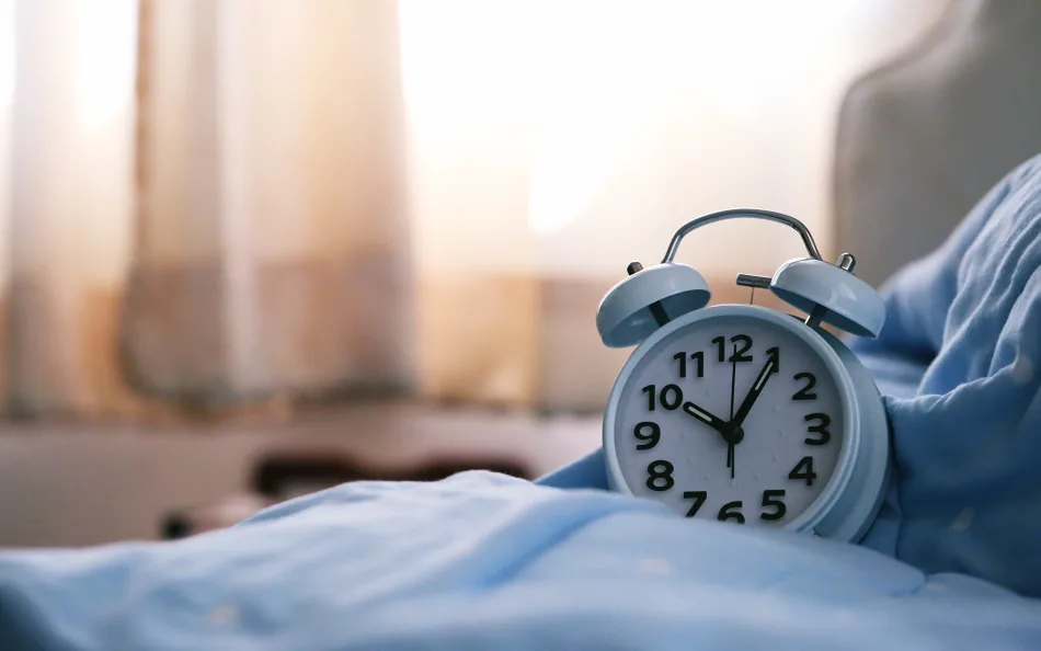 Una vecchia sveglia appoggiata su un letto. Sullo sfondo una finestra illumina la stanza
