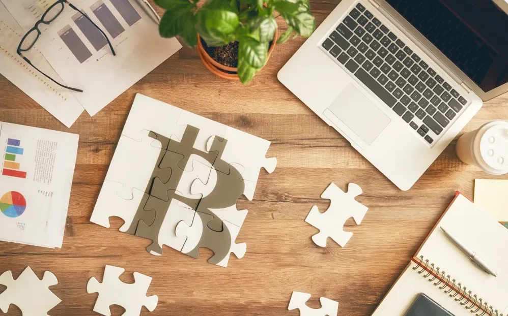 Su una scrivania, vista dall'altro, sono posizionati: un computer, un paio di occhiali, delle slide con grafici, un'agenda con una penna e un puzzle semicomposto che forma il simbolo del Bitcoin