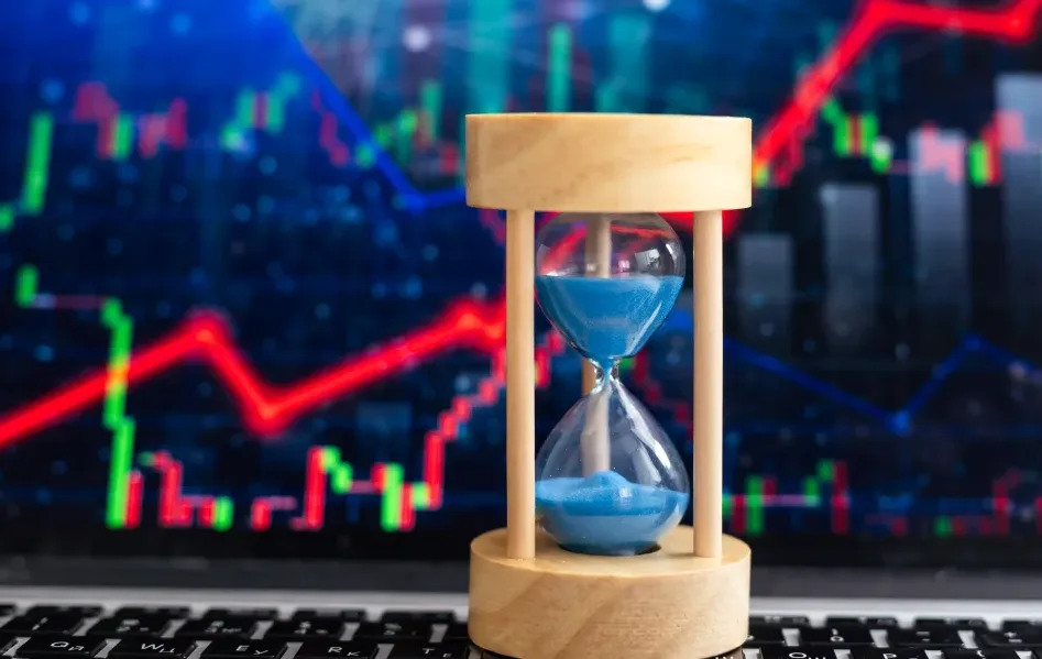 Una clessidra con sabbia blu è posizionata davanti a uno schermo che riproduce le candele rosse e veersi di un grafico di trading