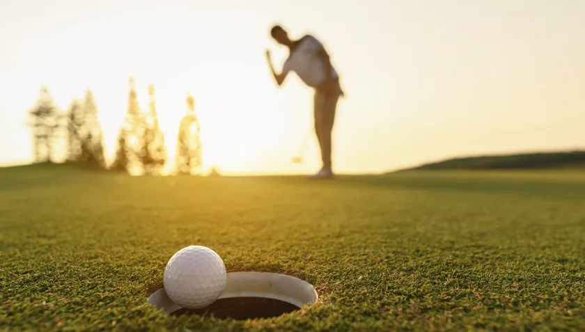 Un golfista, sullo sfondo e in controluce, mette in buca una pallina, inquadrata in primo piano