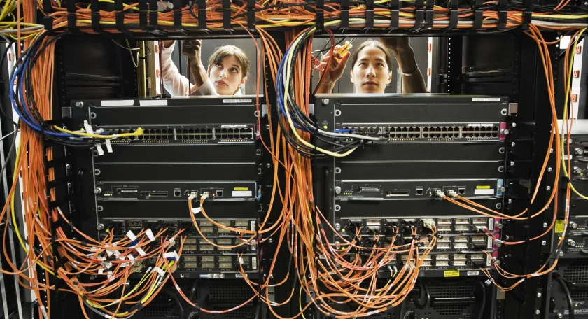 Tra una rete intricata i cavi di un server spuntano le facce di due tecnici al lavoro