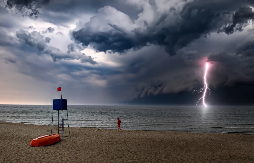 Una spiaggia in un giorno di tempesa. Sullo sfondo, nuvoloni neri e un lampo che cade in mezzo al mare. Sulla spiaggia, il bagnino cammina in riva al mare