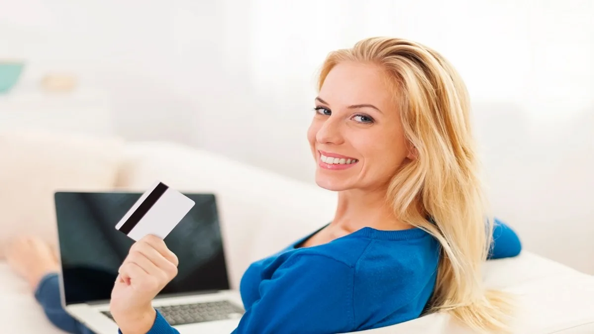 Donna sorridente con carta di credito in mano mentre usa il computer per gestire il conto corrente online