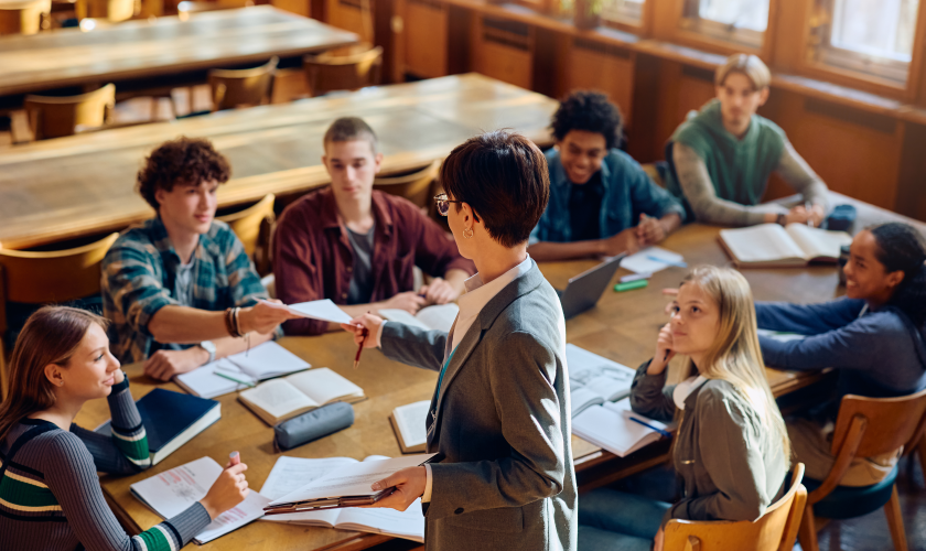 In aula scolastica diversi studendi sono seduti attorno a un tavolo. In piedi, una professoressa ritira il compito di uno dei suoi studenti.