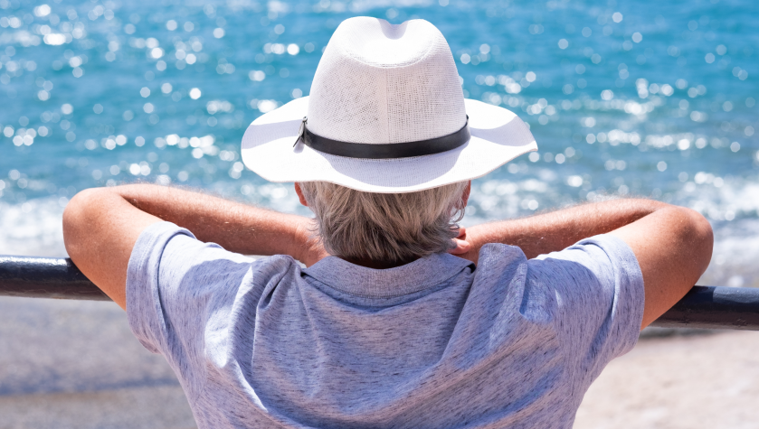 Un pensionato elegante, di spalle e con un cappello, si poggia a un muretto per guardare il mare davanti a sé