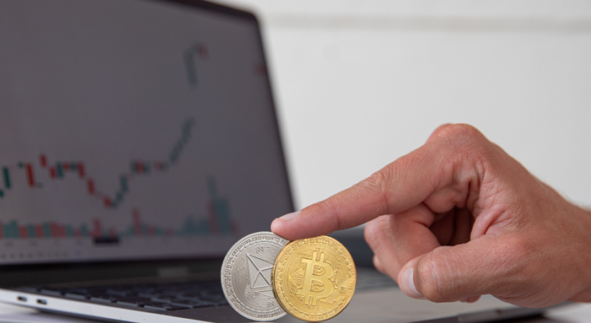 Due monete, un bitcoin e un ethereum vengono sostenute da un dito in verticale davanti allo schermo di un laptop che mostra delle quotazioni di Borsa