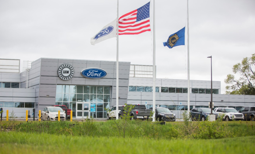 Un'immagine esterna del centro tecnico di addestramento di Ford, negli Usa. In primo piano tre bandiere, una del sindacato Uaw, una della Ford e una degli Usa. Sullo sfondo il centro tecnico con automobili parcheggiate davanti