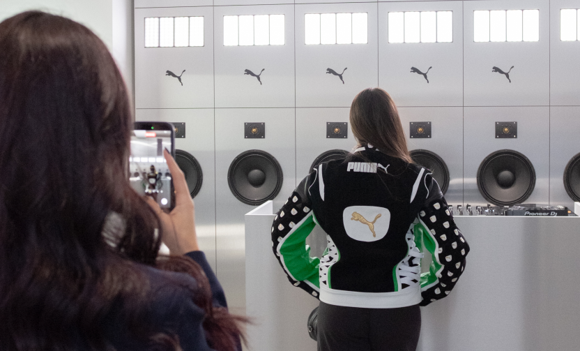 Una ragazza di spalle davanti a un banco con diversi loghi della Puma. Lei stessa indossa uno zainetto Puma. In primo piano, sempre di spalle, un'altra ragazza la sta riprendendo con uno smartphone