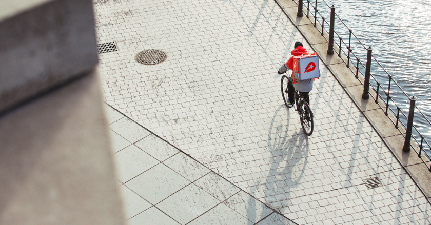 Un addetto alle consegne di Delivery Hero sulla sua bicicletta, con il classico zaino quadrato sulle spalle, sfreccia sul lungo mare di una città non identificabile