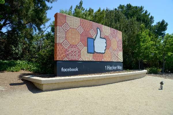 L'immagine mostra l'iconico cartello all'ingresso della sede di Facebook (Meta) a Menlo Park, California, con il noto simbolo del "Mi piace". La segnaletica include l'indirizzo 1 Hacker Way, riconosciuto come il quartier generale dell'azienda. Il contesto verde e il cielo limpido enfatizzano l'ambiente dell'area. Perfetta per contenuti legati a Meta, Facebook, social media, Silicon Valley e aziende tecnologiche.