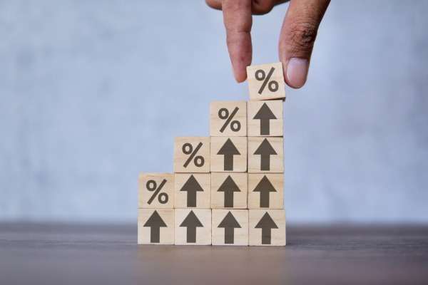 Una mano deposita un quadratino di legno con il simbolo della percentuale su una scala di mattoncini di legno crescente, con frecce verso l'alto e percentuali