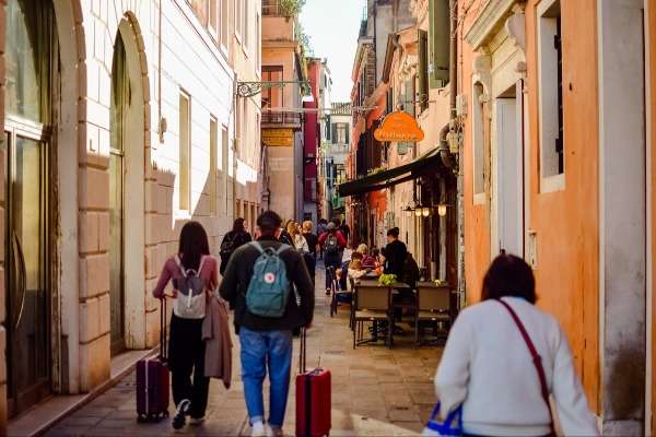 Turisti con valigie in una calle veneziana animata da ristoranti e passanti