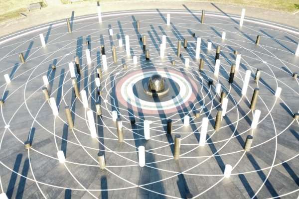 Una piazza con disegni geometrici che si incrociano e numerosi cilindri di diverso colore e altezza che vi proiettano le ombre.