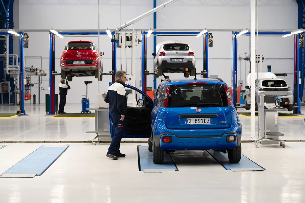 Stabilimento Stellantis con auto sollevate su ponti di controllo e operaio al lavoro su una Fiat Panda blu