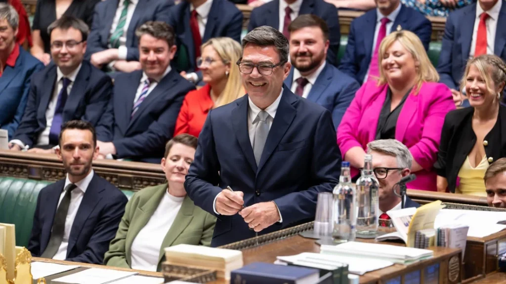 Andy Burnham in piedi tra i banchi della Camera dei Comuni, circondato da deputati laburisti sorridenti