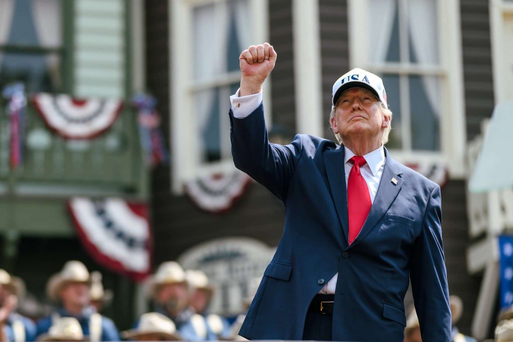 Donald Trump in abito blu e cappello USA alza il pugno destro durante un evento pubblico