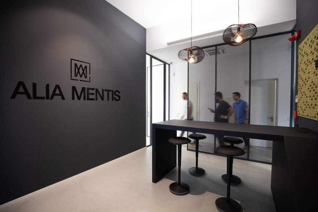 Reception con logo Alia Mentis su parete grigia e area lounge con sgabelli e sospensioni industriali