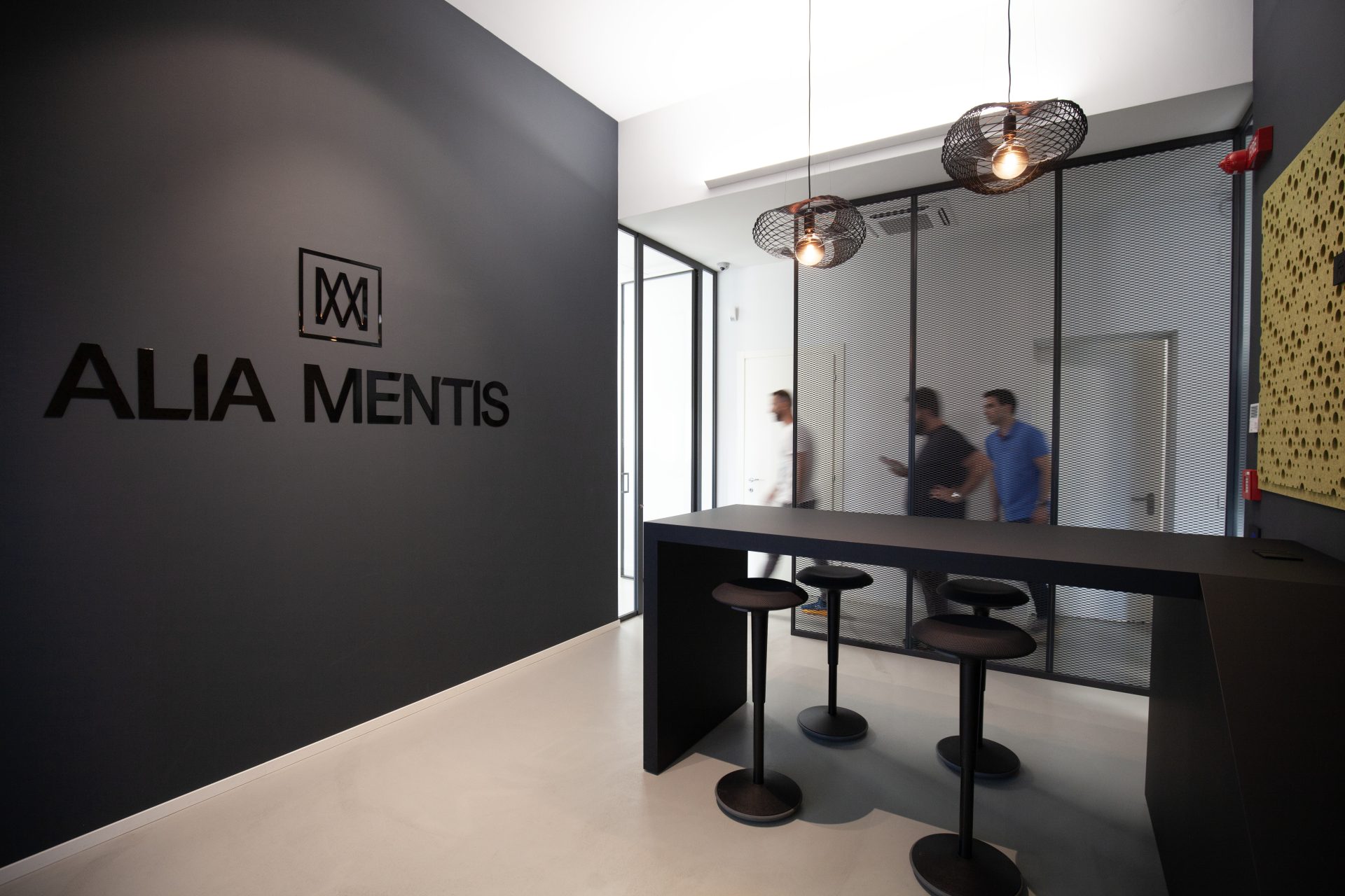 Reception con logo Alia Mentis su parete grigia e area lounge con sgabelli e sospensioni industriali