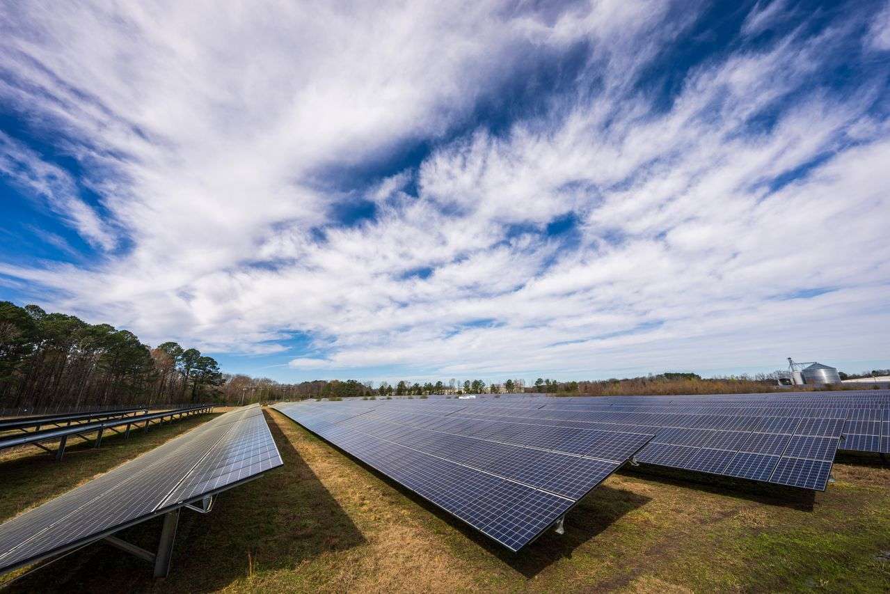 File di pannelli fotovoltaici installati su un ampio terreno agricolo, sotto un cielo con nuvole sparse